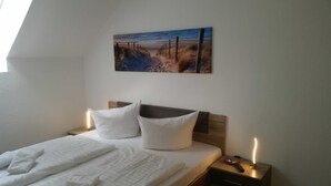 Una camera da letto con un letto, due cuscini bianchi, una testiera in legno, un comodino con una lampada e un quadro raffigurante una spiaggia.