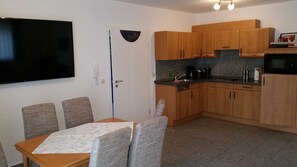 A compact kitchen with wooden cabinets, a microwave, and a coffee maker. There is a dining table with four chairs and a flat-screen TV mounted on the wall.