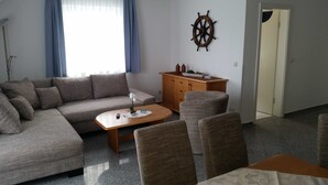 Un soggiorno con un divano, un tavolino da caffè e due poltrone. Si nota un mobile in legno con bottiglie e una pianta sopra. Al muro, una decorazione a forma di timone di nave.