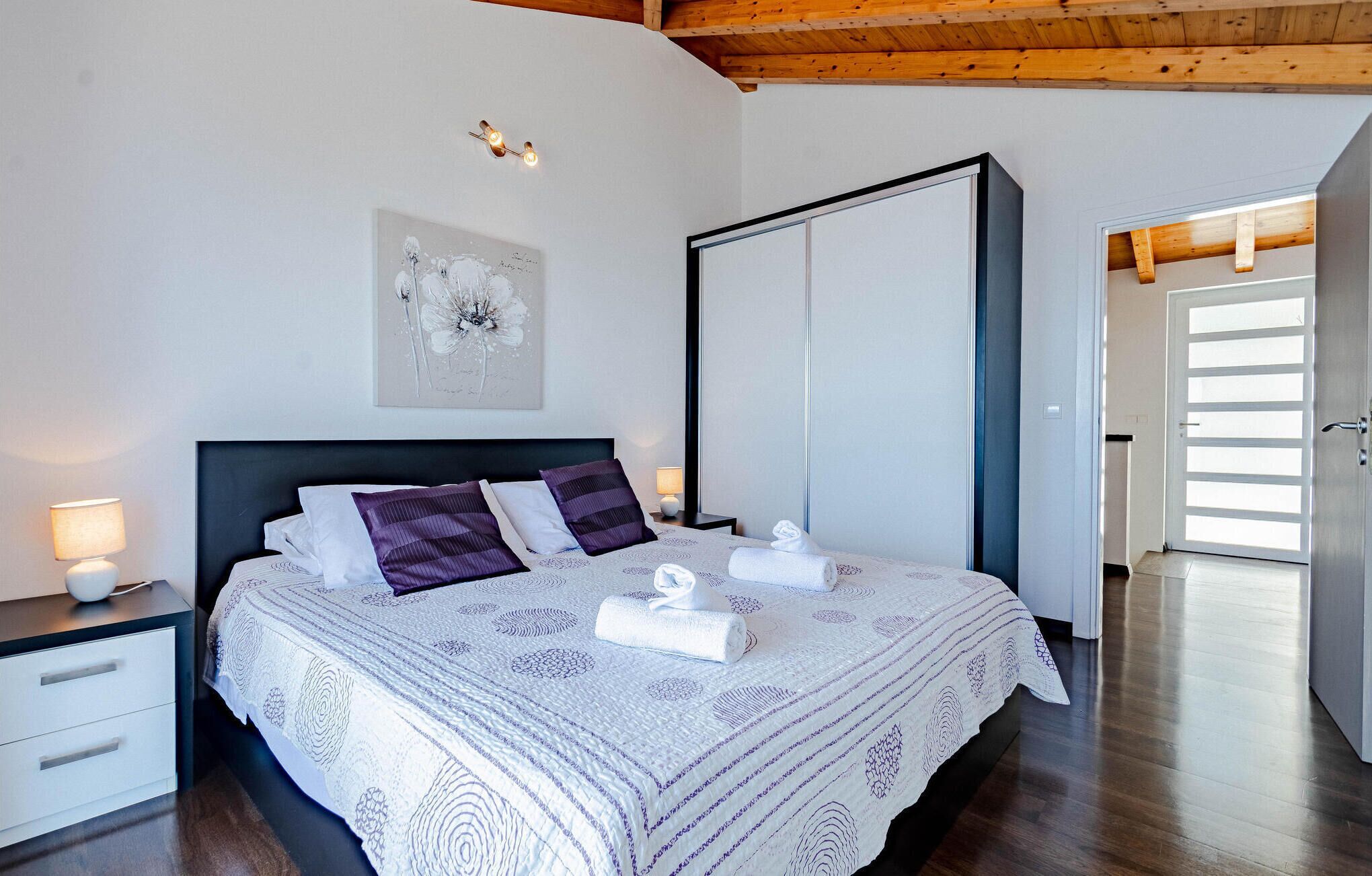 Quarto bem arrumado com uma cama grande, mesa de cabeceira com abajur, uma obra de arte na parede e um guarda-roupa.