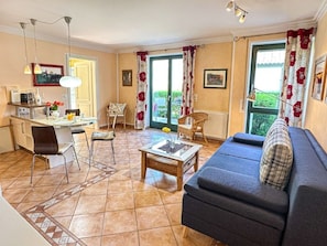 A living room with a sofa, a coffee table, a dining table with chairs, a kitchen area, and a view through a large window. - (210) 2-room apartment - apartment house Villa Laura (Kühlungsborn)