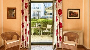Una habitación con vistas a un jardín, dos sillones de mimbre, una mesa y un radiador.