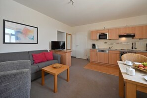 A modern living room with a grey sofa, a wooden coffee table, and a television. Adjacent is a compact kitchen with wooden cabinets, a microwave, and a dining area with a wicker chair and a table set for two.