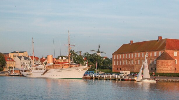 Een jachthaven met verschillende boten afgemeerd, een windmolen op de achtergrond, en een groot, bakstenen gebouw met een rood dak.