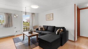 Een woonkamer met een zwarte bank, een houten salontafel en uitzicht op de natuur door een groot raam.
