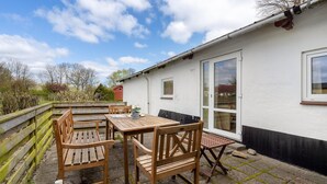 Een terras met houten meubels, een tafel en stoelen, naast een wit gebouw met een deur en ramen.