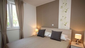 Un letto rifatto con ordine, con copriletto beige e due cuscini, un comodino con una lampada e una finestra con tende.