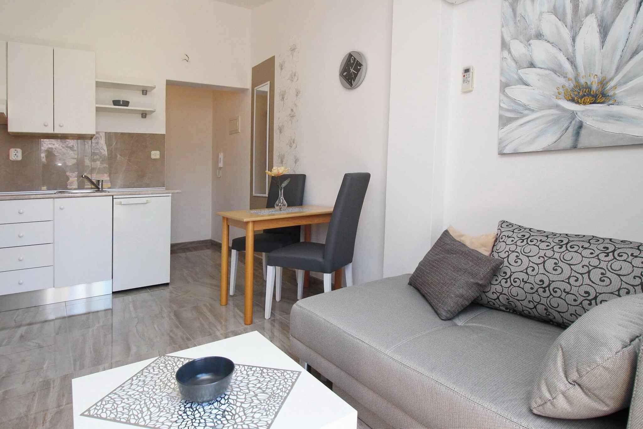A modern living room with a grey sofa, a dining table with a chair, and a kitchen area with white cabinets.