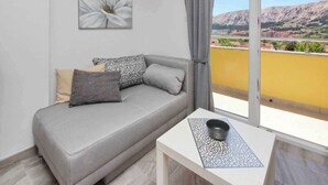 Ett modernt vardagsrum med en grå soffa, mönstrade kuddar, ett litet bord med en skål och ett stort fönster med utsikt över en gul byggnad och berg.