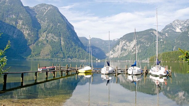 A calm lake with several sailboats docked at a wooden pier, surrounded by mountains.