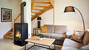 A modern living room with a wood-burning stove, a grey sofa, a coffee table, and a staircase with exposed wooden beams.