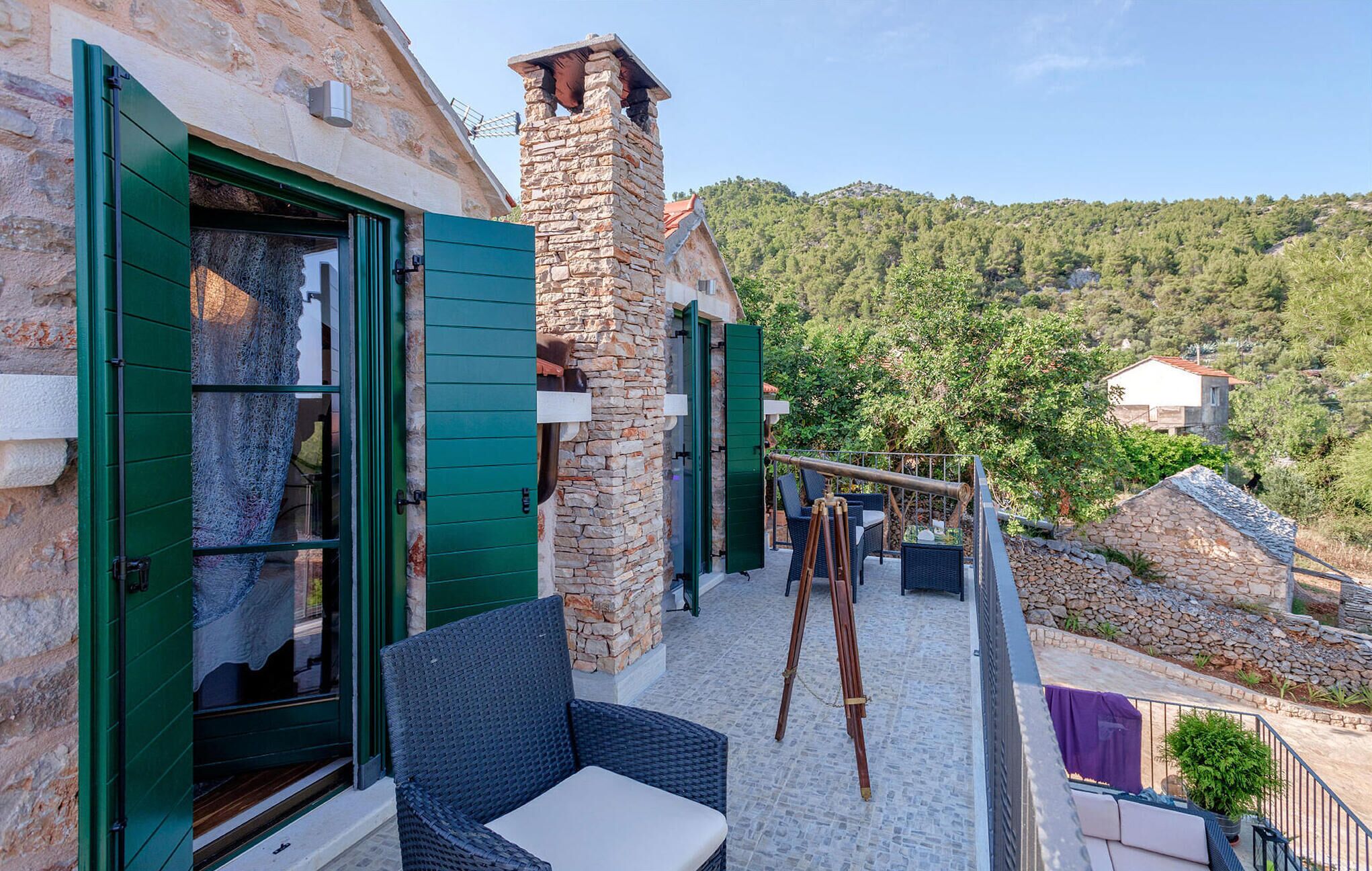 Ein Steinhaus mit grünen Fensterläden, einem Balkon mit Korbmöbeln und Blick auf einen bewaldeten Hügel.