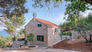 A stone house with a red-tiled roof, a satellite dish, and a small garden area with trees and shrubs.