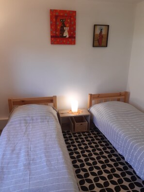 A small, simple room with two single beds, a nightstand with a lamp, and two framed pictures on the wall.