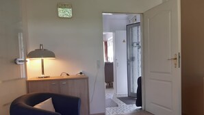 Un corridoio con una poltrona blu, una scrivania con una lampada, una sveglia e una porta che dà su un'altra stanza.