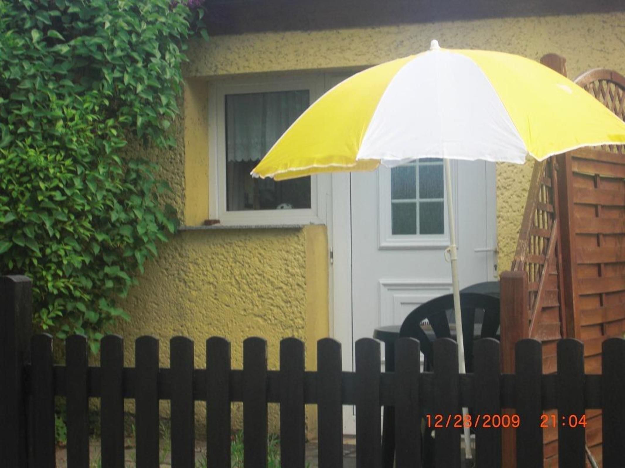 Un paraguas amarillo y blanco frente a una casa con puerta y ventana blancas, rodeada por una valla de madera.