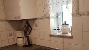Une cuisine équipée d’un évier à deux bacs, de meubles blancs, d’une machine à café et d’une petite fenêtre agrémentée d’un rideau décoratif.