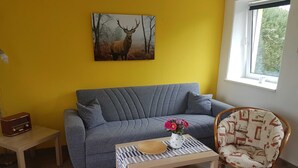 Un salon avec un canapé gris, une petite table sur laquelle est posé un vase de fleurs, un fauteuil en osier et un tableau représentant un cerf accroché au mur.