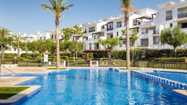 A swimming pool surrounded by white buildings with balconies, palm trees, and a pergola.