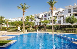 A swimming pool surrounded by white buildings with balconies, palm trees, and a pergola.