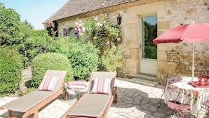 Un bâtiment en pierre doté d’un patio carrelé, de deux chaises longues avec des coussins à rayures, d’une table avec un parasol rouge et d’un pot de fleurs orné de fleurs roses.