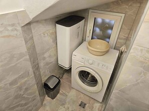 A compact bathroom with a washing machine, a small trash bin, and a glass-enclosed toilet.