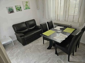 Et spiseområde med en sort lædersofa, et spisebord med en hvid blondeopsætning og et flisegulv.