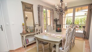 Un comedor con candelabro, sillas ornamentadas, una mesa con mantel floral y un amplio ventanal con cortinas.