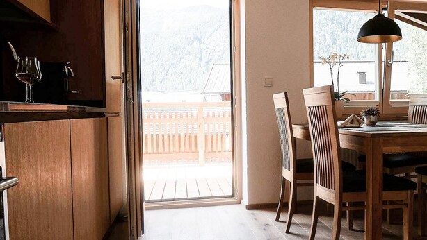 Un comedor con mesa y sillas de madera, una lámpara colgante y vistas a una terraza de madera y árboles al exterior.
