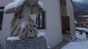 Una estatua de piedra de un águila con nieve en las alas y las patas, ubicada sobre un pedestal frente a un edificio con techo inclinado y una ventana.