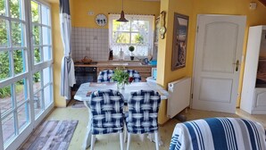 A dining area with a table and chairs, a kitchen in the background, and a large window with curtains.