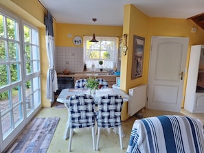 A dining area with a table and chairs, a kitchen in the background, and a large window with curtains.