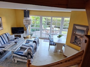 A living room with a sofa, coffee table, and TV, a dining area with chairs, and a large window with a view of greenery.