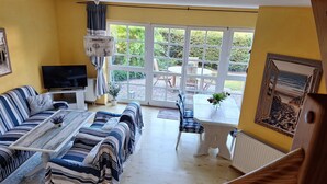 A living room with a sofa, coffee table, and TV, a dining area with chairs, and a large window with a view of greenery.