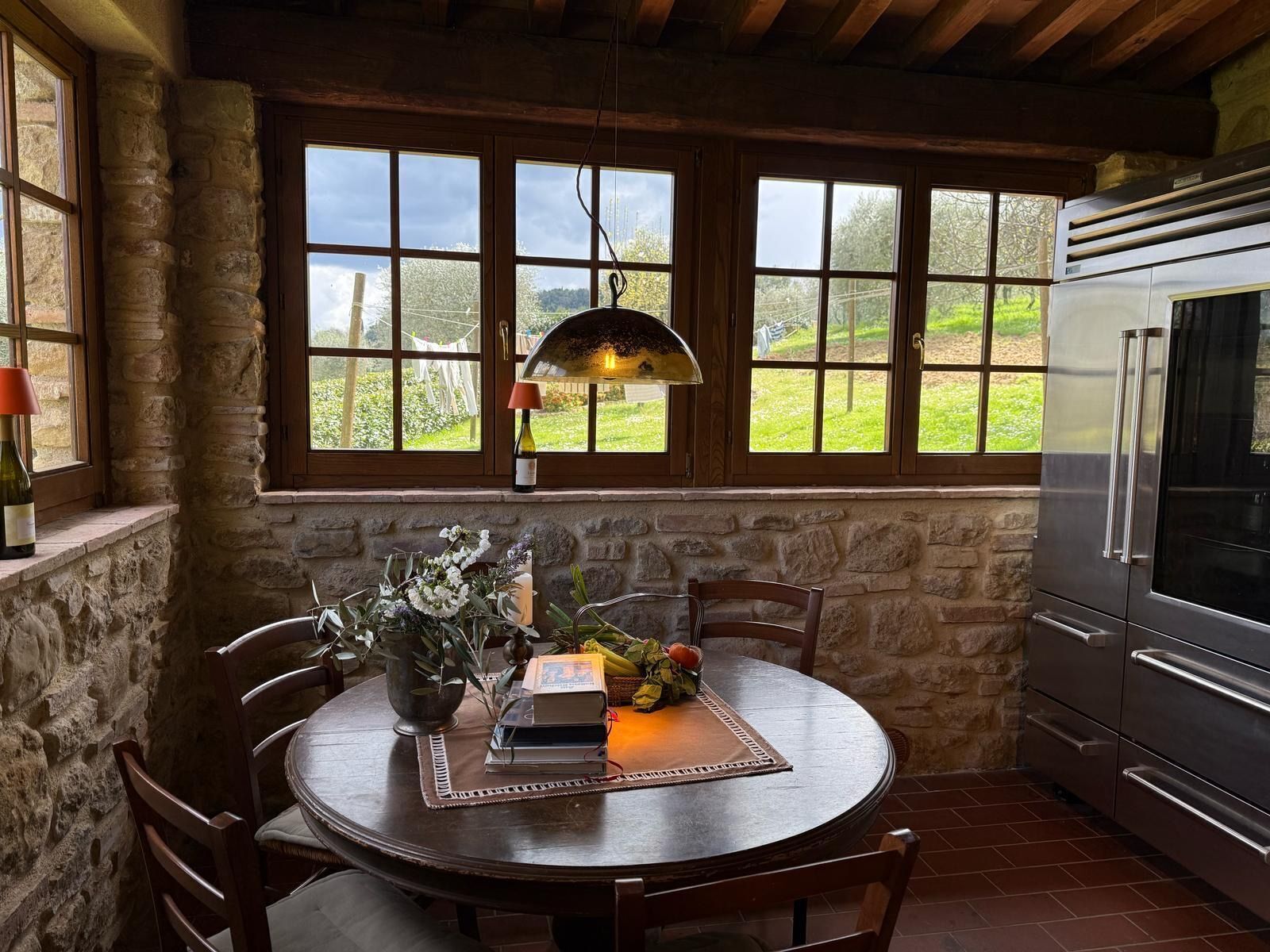 Una cocina rústica con paredes de piedra, una mesa redonda de madera con sillas, un ventanal con vistas a la vegetación y un refrigerador moderno de acero inoxidable.