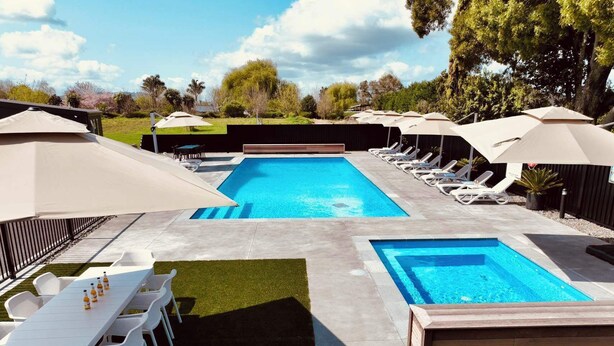 A swimming pool with two separate sections, surrounded by lounge chairs and umbrellas, with a table and chairs nearby. - Podium Lodge (Cambridge)