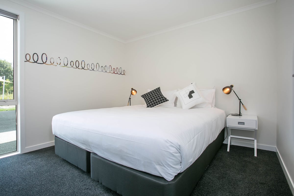 A neatly arranged bedroom with a large bed, two bedside tables with lamps, a wall art piece, and a view through a door.