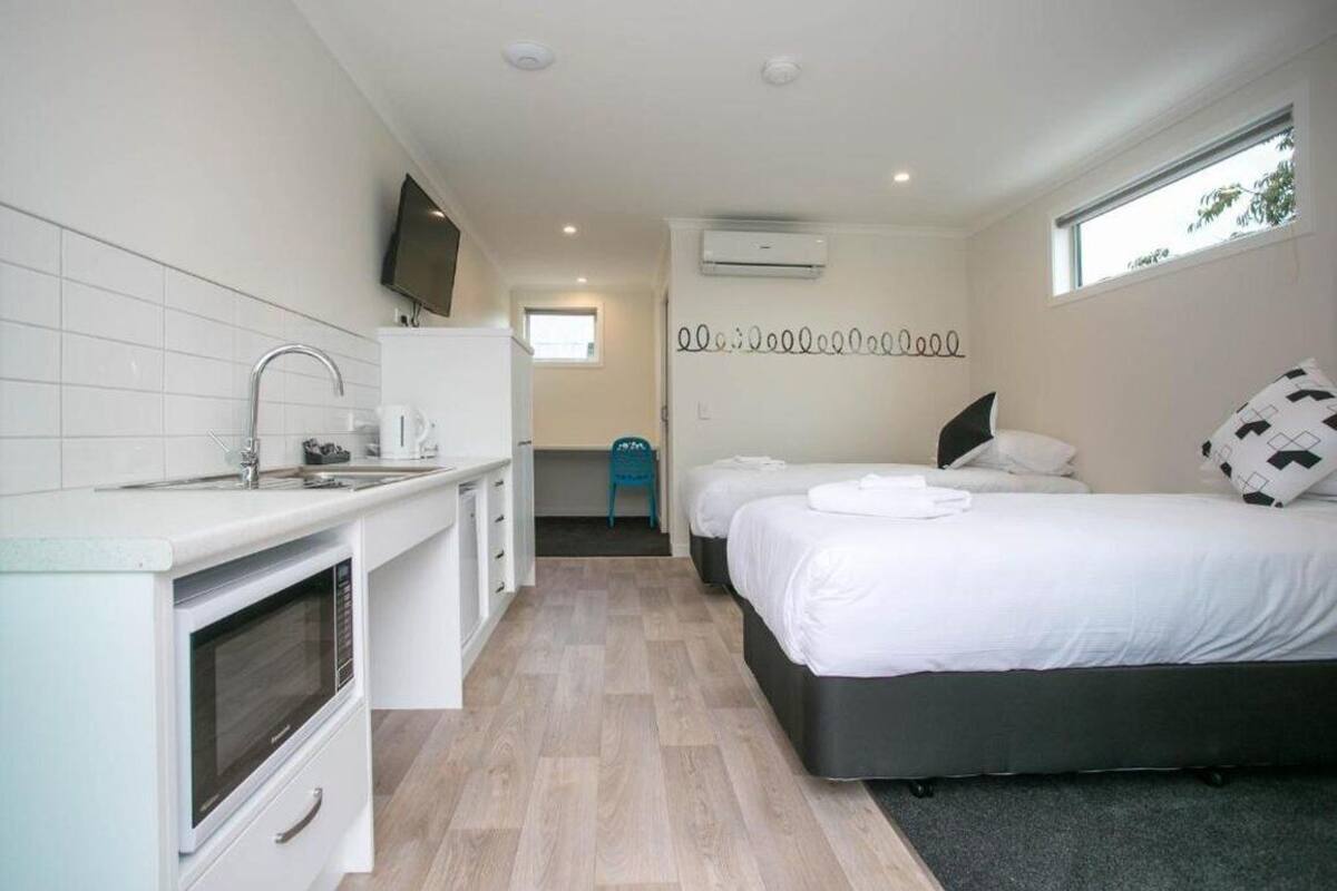 A compact hotel room with a kitchenette, a bed with white bedding, a small bathroom, and a window providing natural light.
