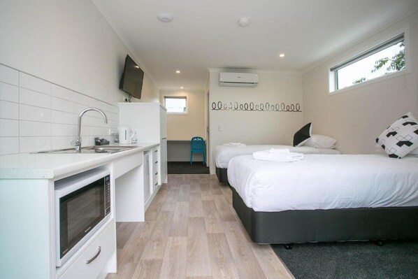 A compact hotel room with a kitchenette, a bed with white bedding, a small bathroom, and a window providing natural light.