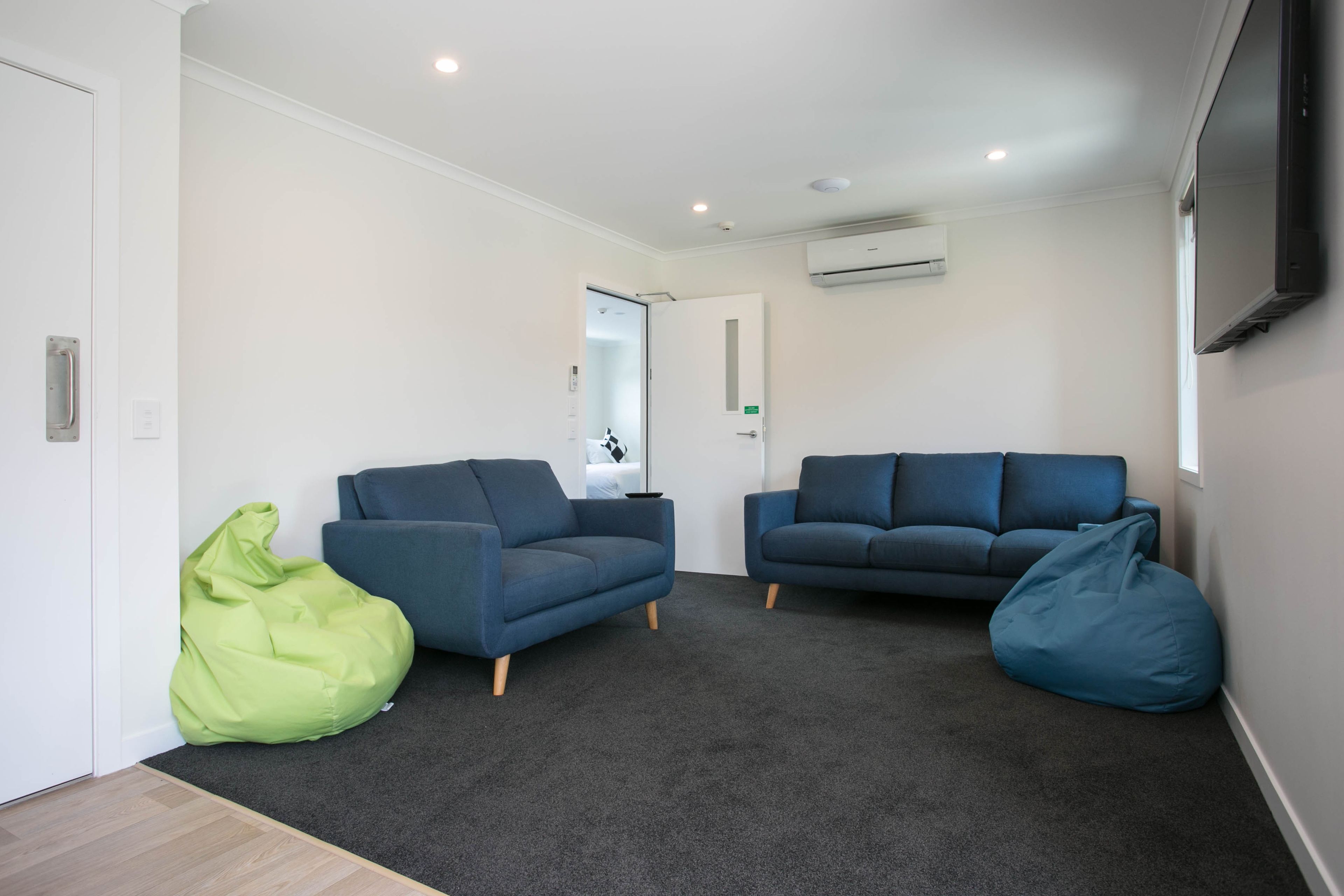 A modern living room with two sofas and two bean bags, carpeted floor, white walls, and an air conditioning unit.