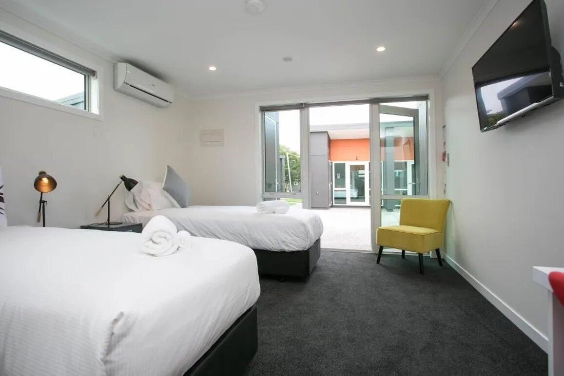 A hotel room with two beds, a flat-screen TV, a yellow chair, and a sliding glass door leading to an outdoor area.