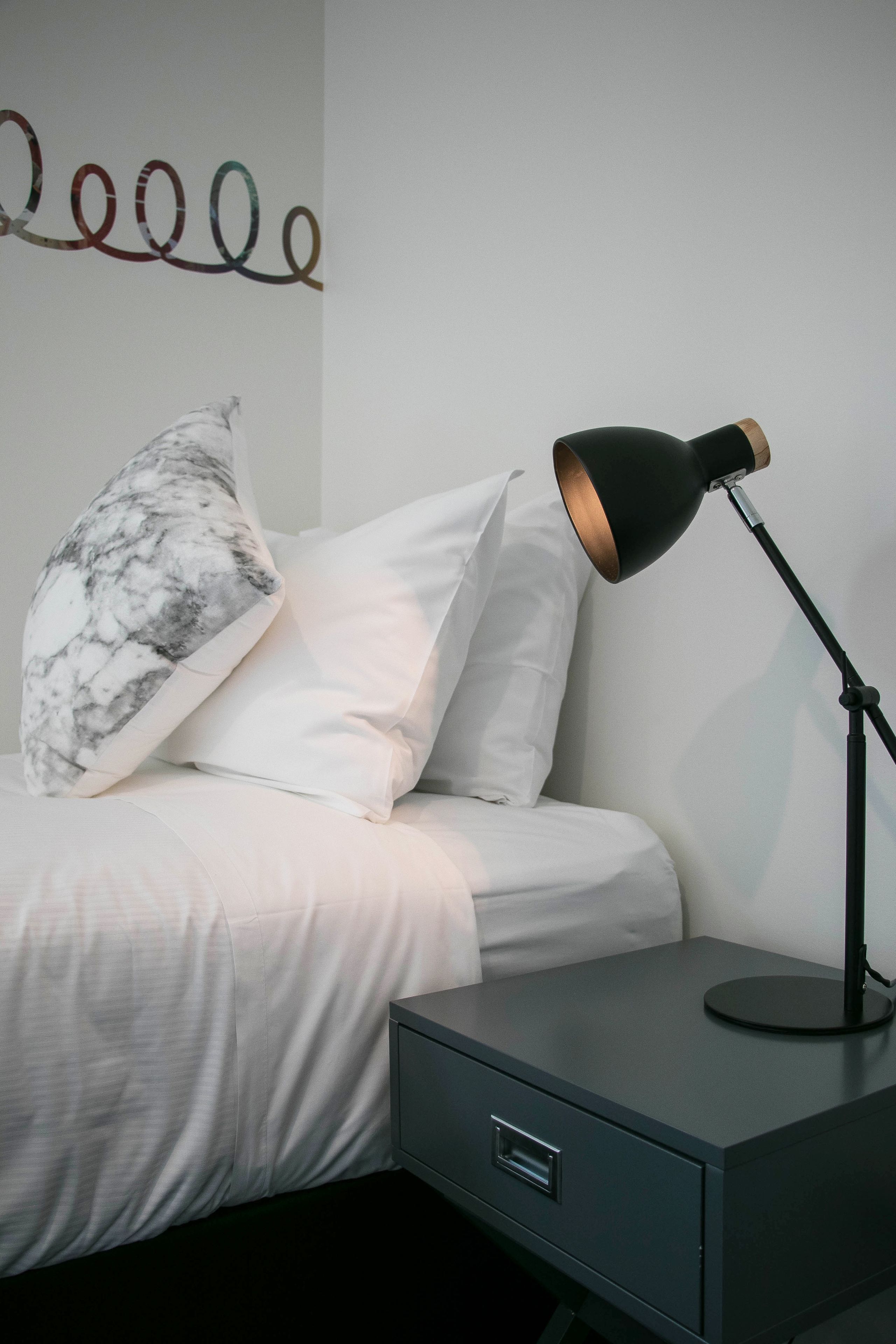A neatly made bed with white bedding and a marble-patterned pillow, a black desk lamp on a bedside table, and a small drawer unit.