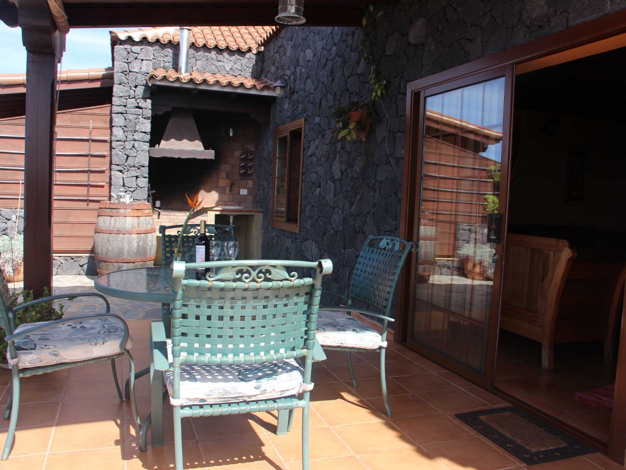 A patio with a glass table and two chairs, a stone wall, and a view through a sliding glass door.
