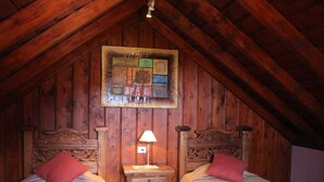 Ein gemütliches Schlafzimmer im Dachgeschoss mit Holzbalken, zwei Betten mit roten Kissen, einem Nachttisch mit Lampe und einem gerahmten Kunstwerk an der Wand.