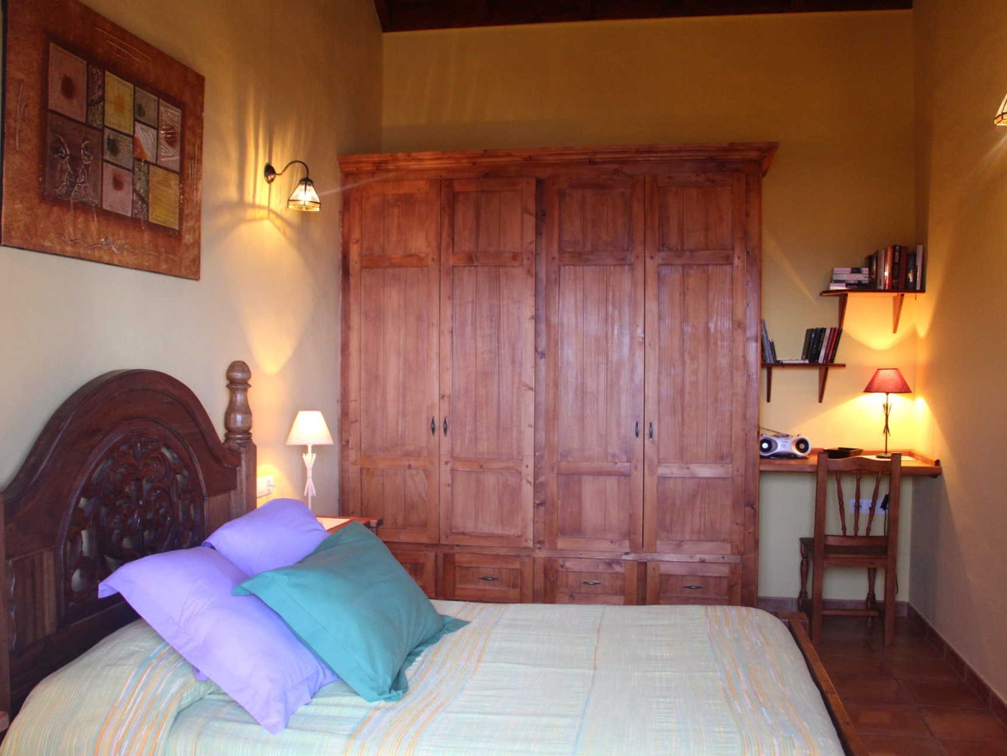 Quarto com um grande armário de madeira, uma cama com cabeceira, duas luminárias de cabeceira e uma pequena mesa com uma luminária e livros.