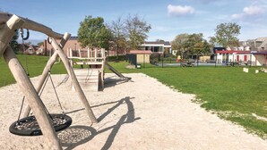 A playground with swings and a slide, surrounded by a fenced area and a grassy field.