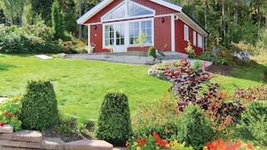 Eine rote Holzhütte mit großer Glasscheibe, umgeben von einem gepflegten Garten mit verschiedenen Sträuchern und Blumen.