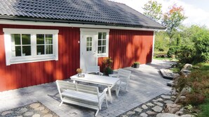 Ein rotes Gebäude mit weißer Tür und Fenstern, eine gepflasterte Terrasse mit Tisch und Stühlen sowie ein Garten mit Steinen und Pflanzen.