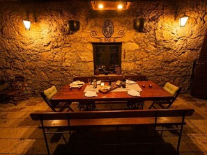 Un coin repas aux murs de pierre, avec une table en bois dressée pour quatre personnes, des bancs et des lanternes fixées au mur.