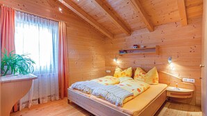 A wooden cabin bedroom with a bed, curtains, a bedside table, a shelf, and a window with a plant. - Ferienwohnung - Hoch Pustertal (Kartitsch)